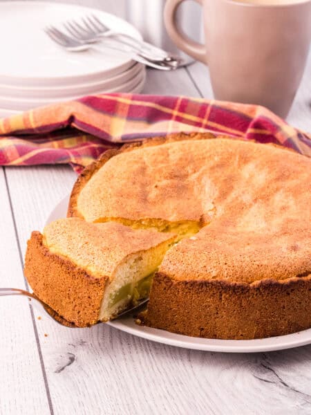 whole golden brown rhubarb cake with on slice slightly elevated on a cake shovel. Cups, plates and forks are in the background.