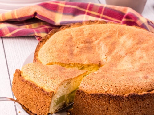 whole golden brown rhubarb cake with on slice slightly elevated on a cake shovel. Cups, plates and forks are in the background.