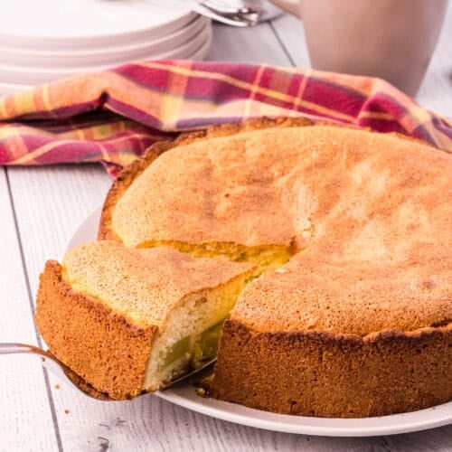 whole golden brown rhubarb cake with on slice slightly elevated on a cake shovel. Cups, plates and forks are in the background.