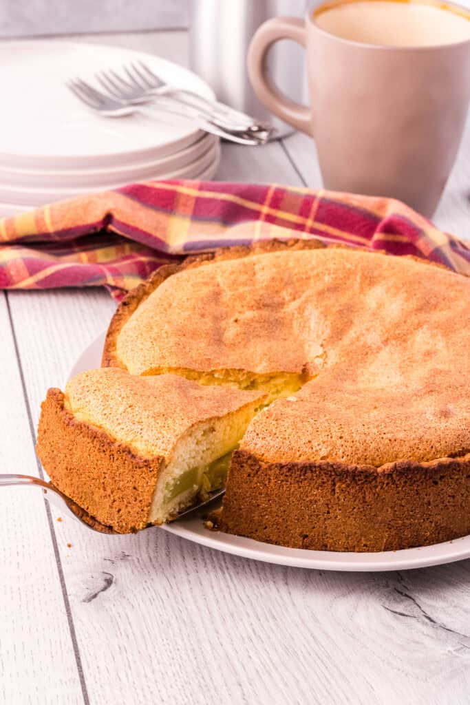 whole golden brown rhubarb cake with on slice slightly elevated on a cake shovel. Cups, plates and forks are in the background.
