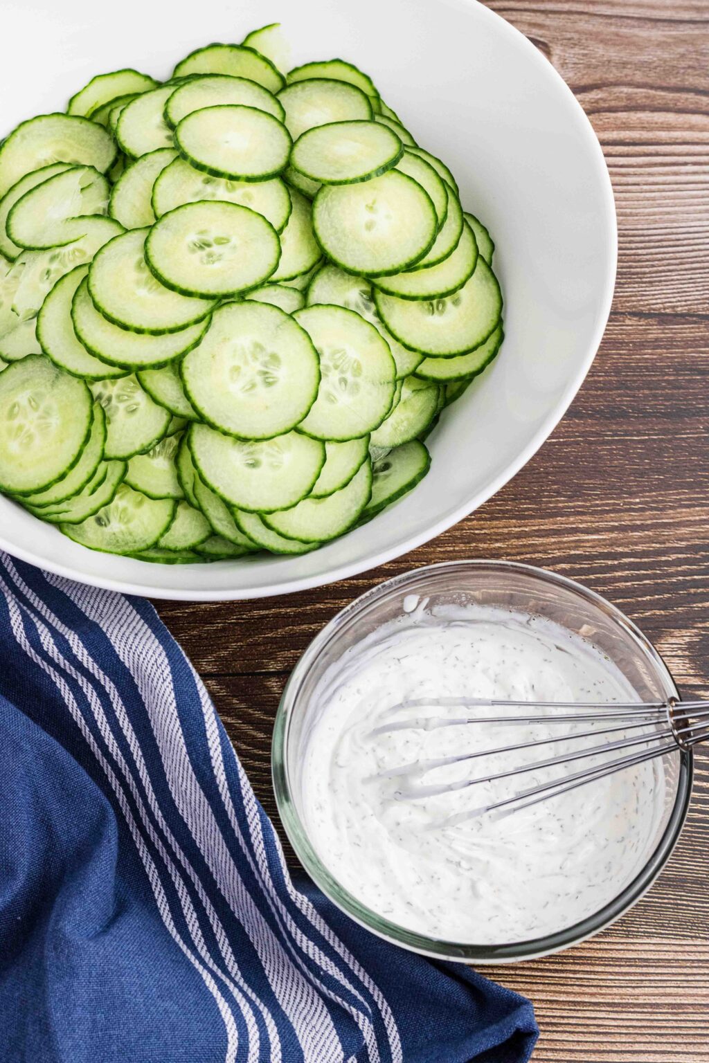 plain cucumbers in white salad bowl with finished dressing in front of cucumbers. Dressing sits in a glass bowl with the whisk still in it