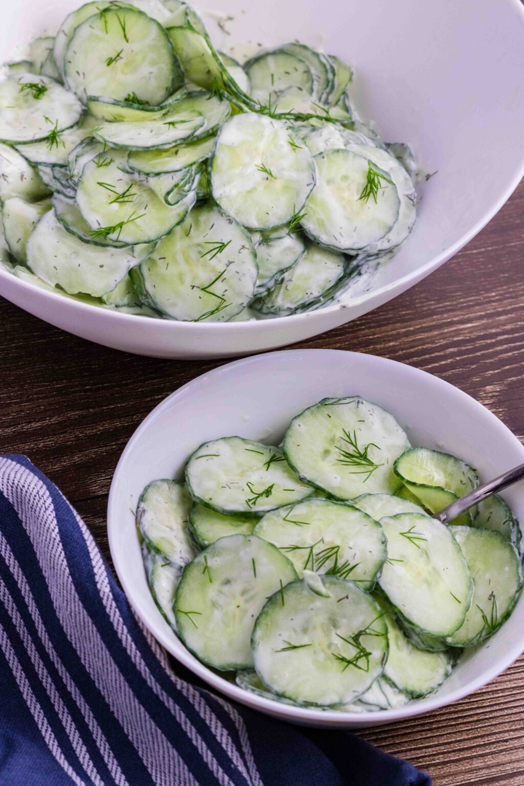 small white salad bowl with cucumber salad in the front. Blue town laying on the left side and main salad with cucumber salad in the back.