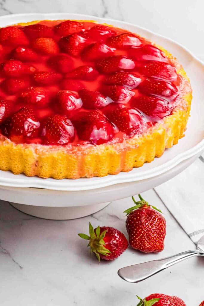 strawberry sponge cake with red glazed strawberries on a white cake stand.