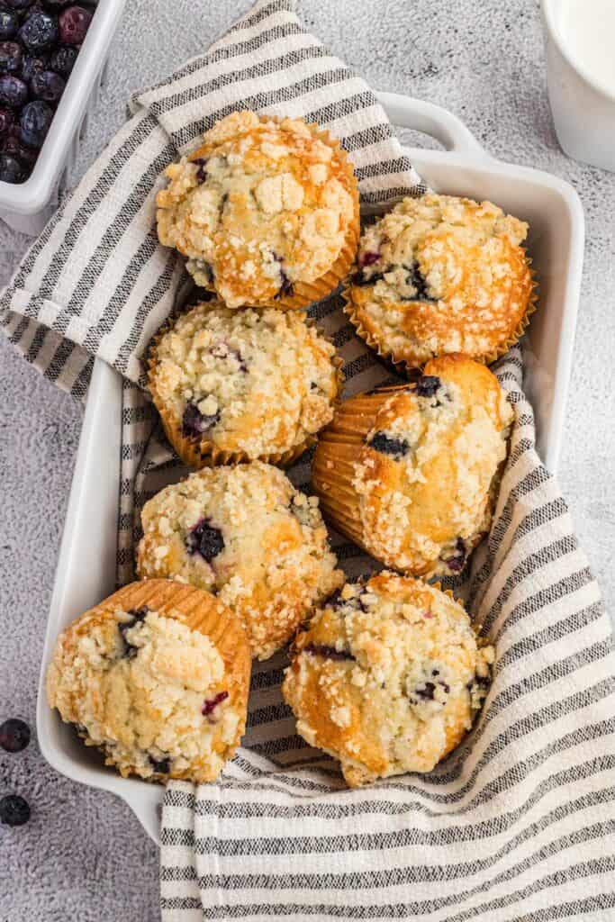 8 streusel topped blueberry muffins in a striped towel in a white loaf form.