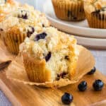 Three blueberry muffins on a wood board. The front muffin has a piece bitten off.