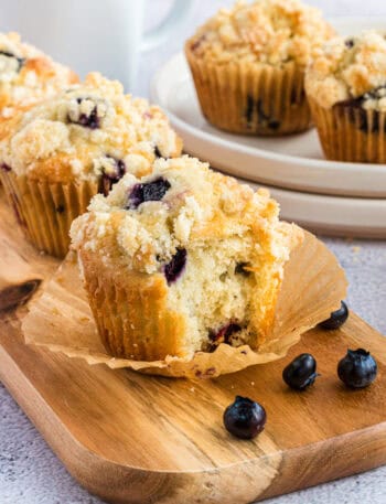 Three blueberry muffins on a wood board. The front muffin has a piece bitten off.