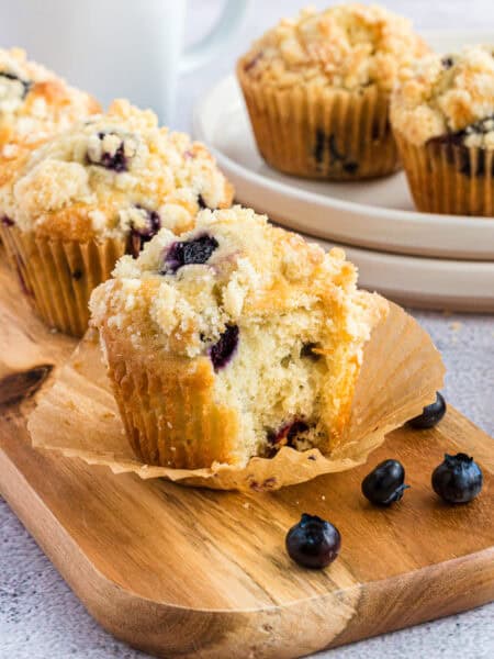 Three blueberry muffins on a wood board. The front muffin has a piece bitten off.