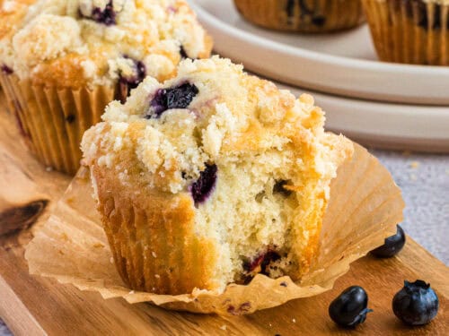 Three blueberry muffins on a wood board. The front muffin has a piece bitten off.