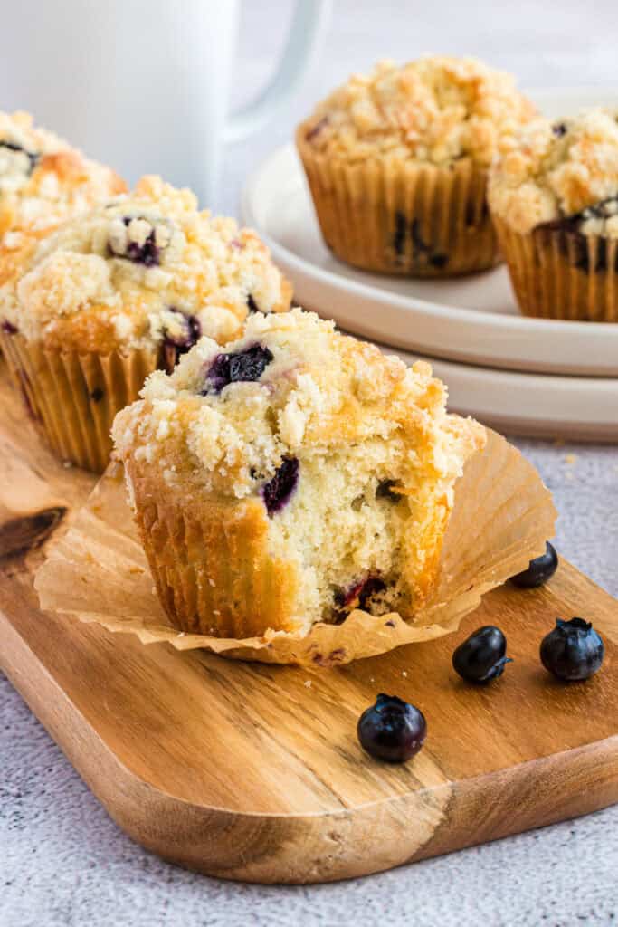Three blueberry muffins on a wood board. The front muffin has a piece bitten off.