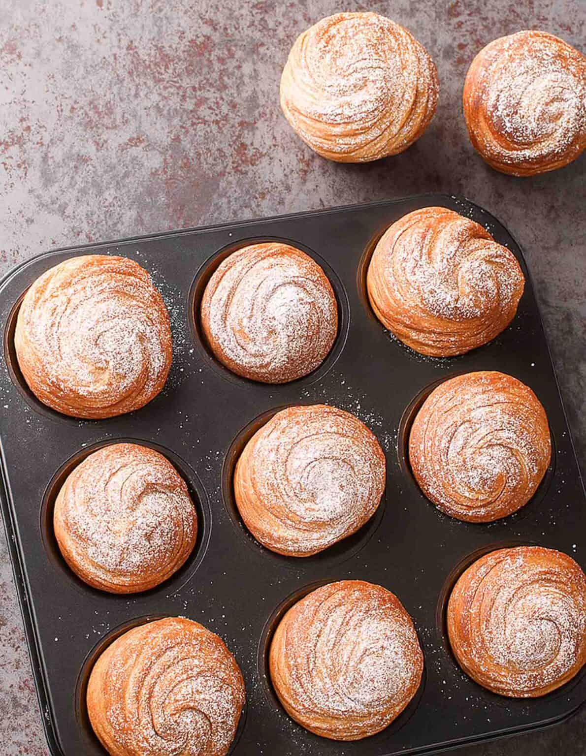 9 cruffins still in baking tin with 2 cruffins on the top outside. All powdered with powder sugar