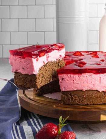 Elevated slice of strawberry mousse cake with bottom chocolate sponge followed by strawberry mousse and finished with layered strawberries glazed with red strawberry jello. Placed on a dark wood round cake stand.