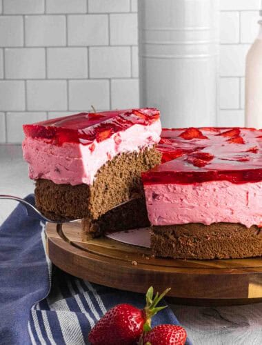 Elevated slice of strawberry mousse cake with bottom chocolate sponge followed by strawberry mousse and finished with layered strawberries glazed with red strawberry jello. Placed on a dark wood round cake stand.
