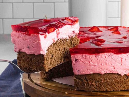 Elevated slice of strawberry mousse cake with bottom chocolate sponge followed by strawberry mousse and finished with layered strawberries glazed with red strawberry jello. Placed on a dark wood round cake stand.