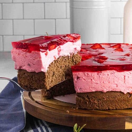 Elevated slice of strawberry mousse cake with bottom chocolate sponge followed by strawberry mousse and finished with layered strawberries glazed with red strawberry jello. Placed on a dark wood round cake stand.