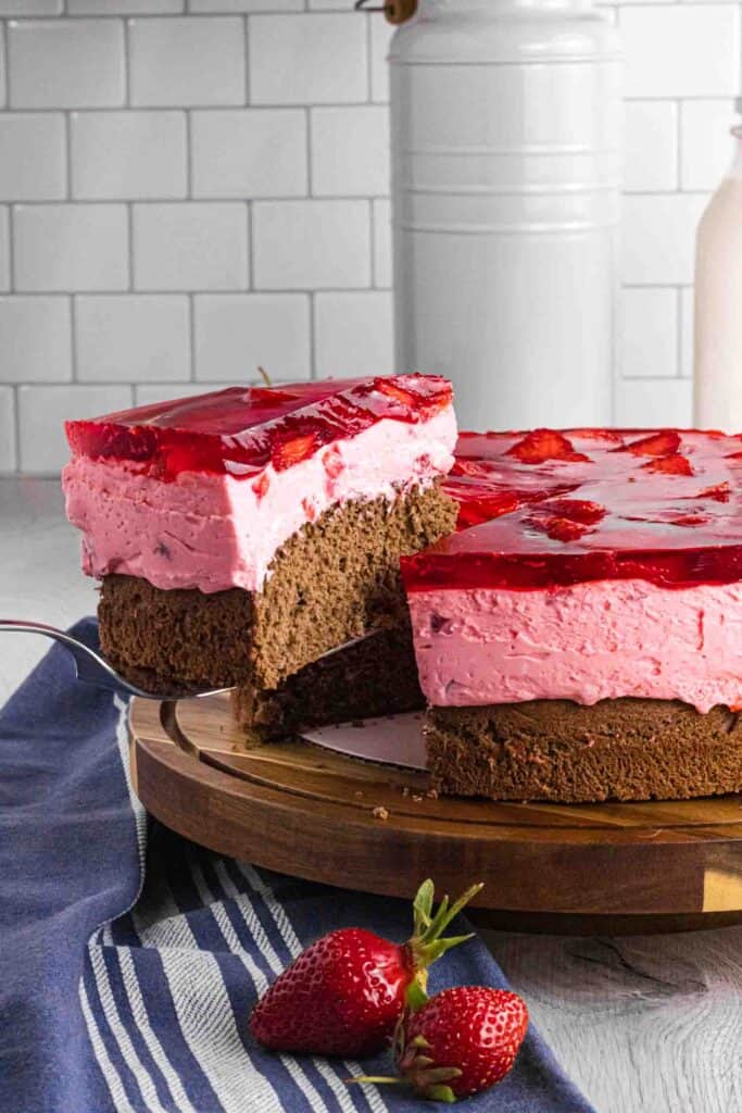 Elevated slice of strawberry mousse cake with bottom chocolate sponge followed by strawberry mousse and finished with layered strawberries glazed with red strawberry jello. Placed on a dark wood round cake stand.