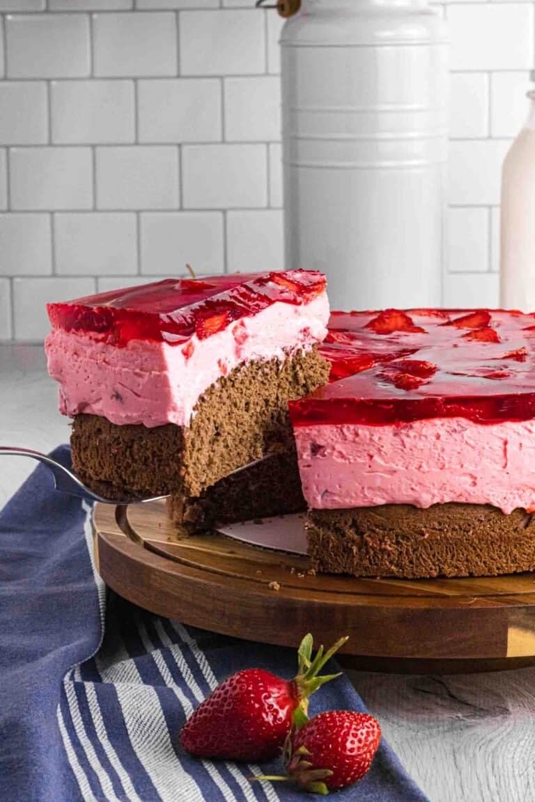 Elevated slice of strawberry mousse cake with bottom chocolate sponge followed by strawberry mousse and finished with layered strawberries glazed with red strawberry jello. Placed on a dark wood round cake stand.