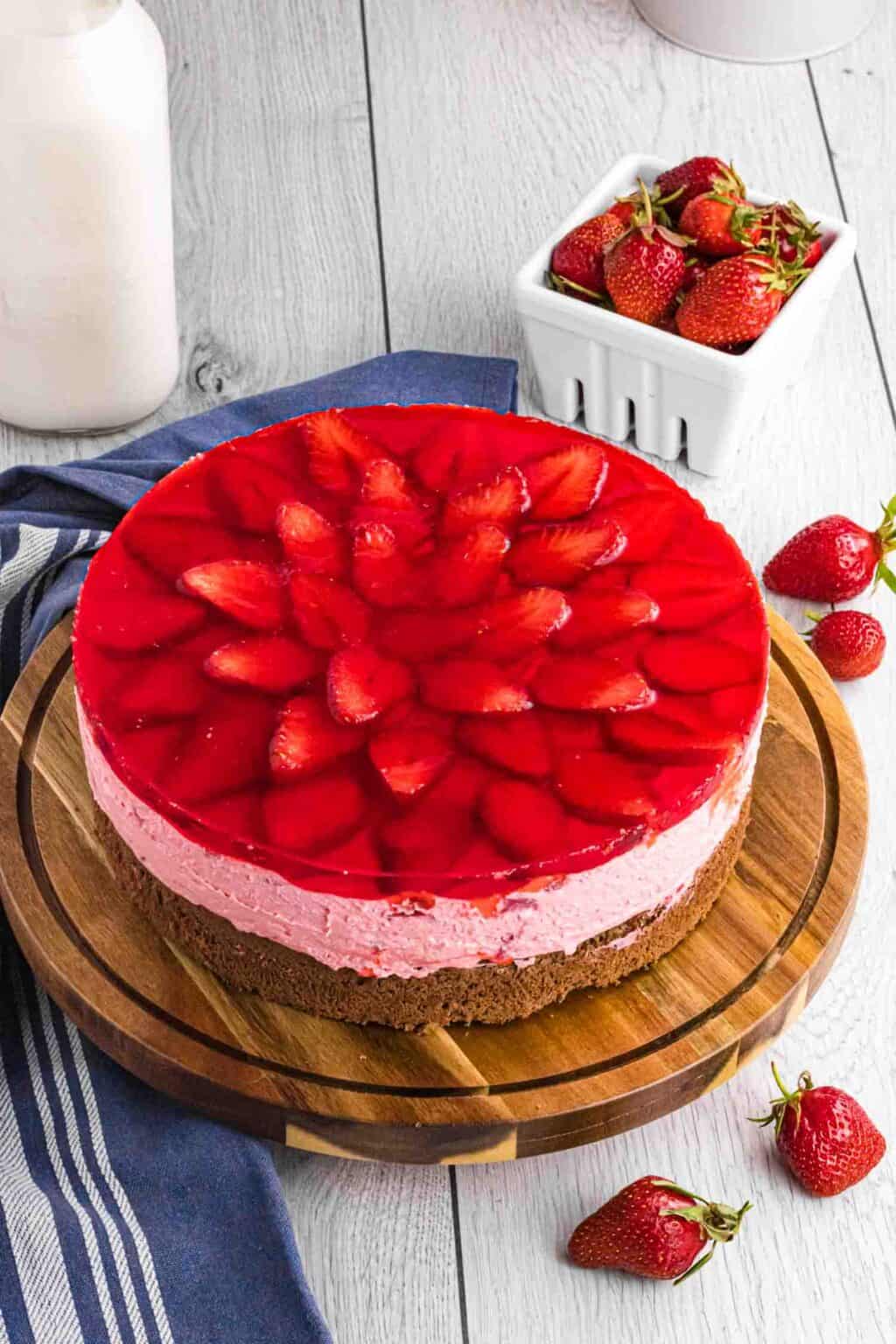 strawberry mousse cake shown as a whole from top on a round wooden cake stand with a white strawberry basket full of berries standing behind on the right of the cake.