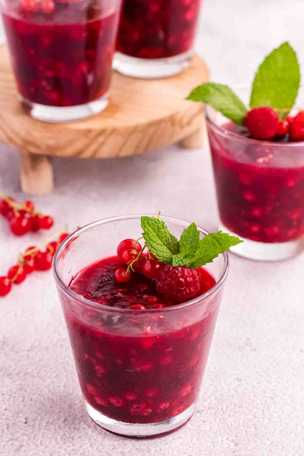 4 glasses of Rote Grütze decorated with raspberries, currants and mint leaves. One glass in the front and 3 more in the background.