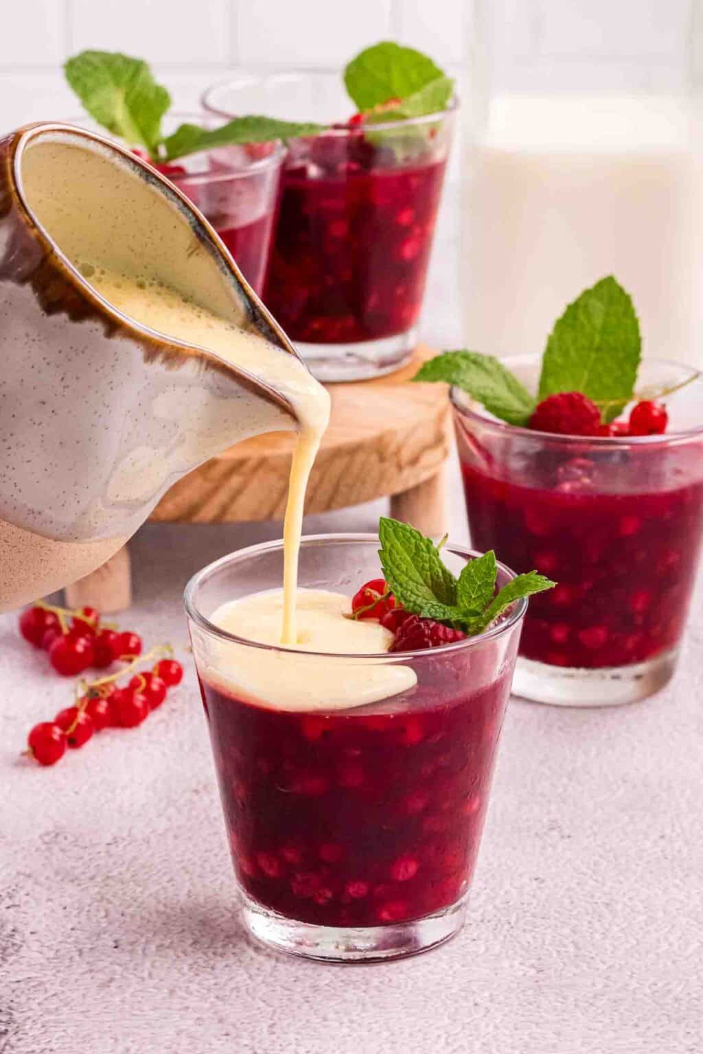 Rote Grütze in a glasses decorated with raspberries, currants and a mint leaf. Vanilla sauce is being poured on top of the front glass.