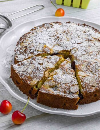 Powdered cherry cake on a white plate. Has two slices already cut and sticking out a bit. Decorated with a couple of cherries