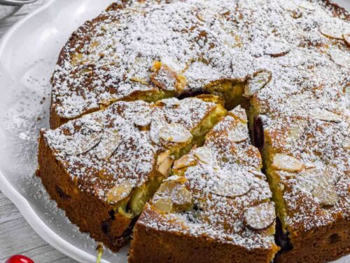 Powdered cherry cake on a white plate. Has two slices already cut and sticking out a bit. Decorated with a couple of cherries