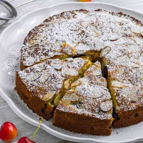 Powdered cherry cake on a white plate. Has two slices already cut and sticking out a bit. Decorated with a couple of cherries