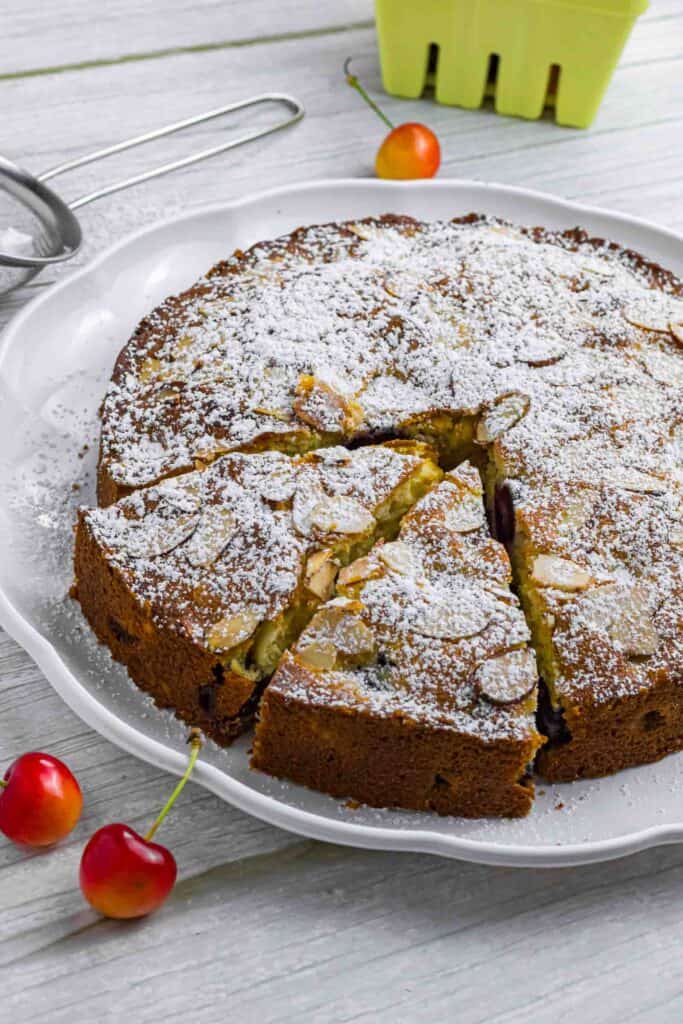 Powdered cherry cake on a white plate. Has two slices already cut and sticking out a bit. Decorated with a couple of cherries