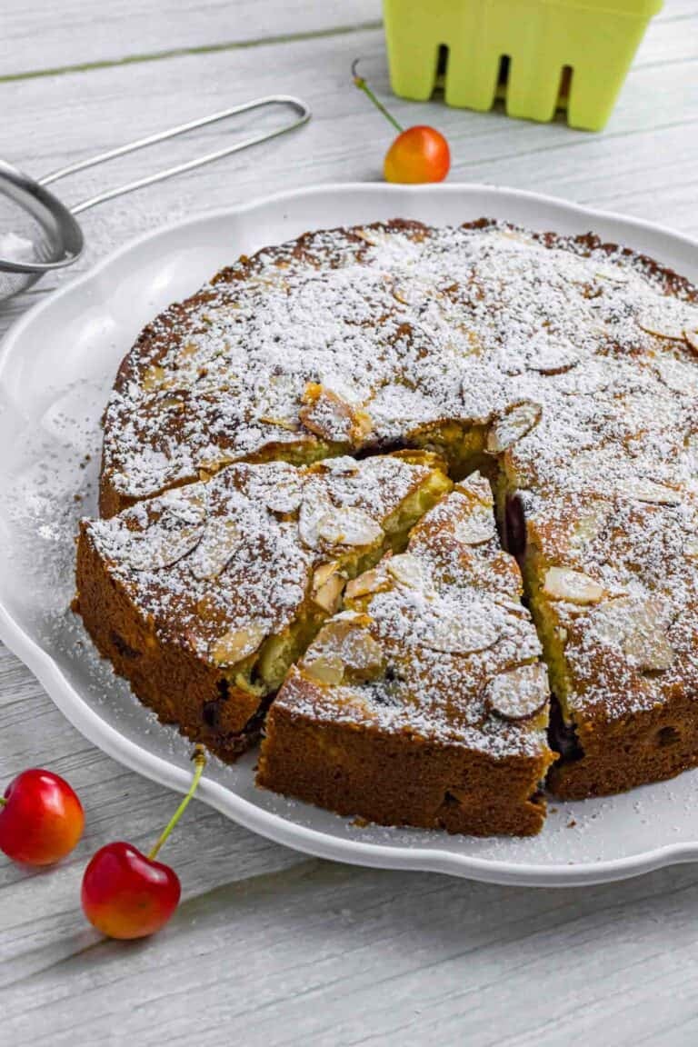 Powdered cherry cake on a white plate. Has two slices already cut and sticking out a bit. Decorated with a couple of cherries