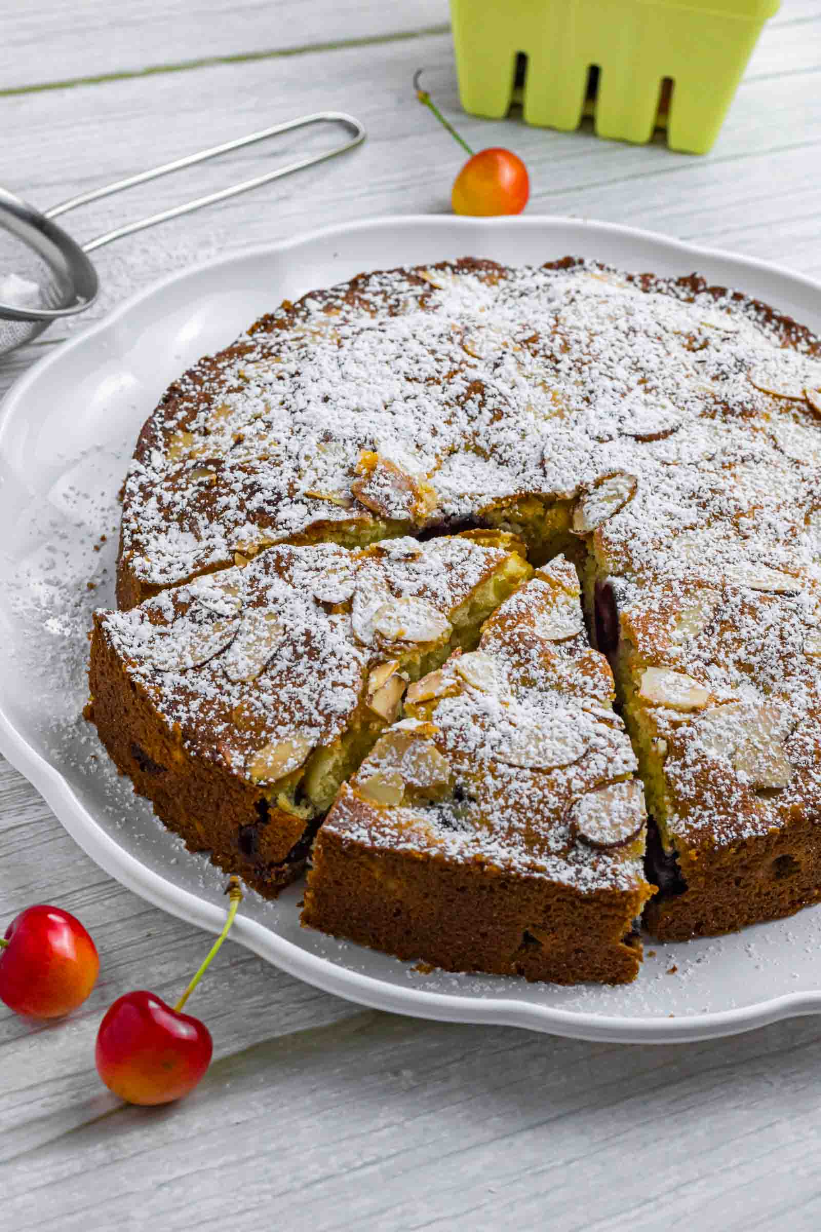 Powdered cherry cake on a white plate. Has two slices already cut and sticking out a bit. Decorated with a couple of cherries