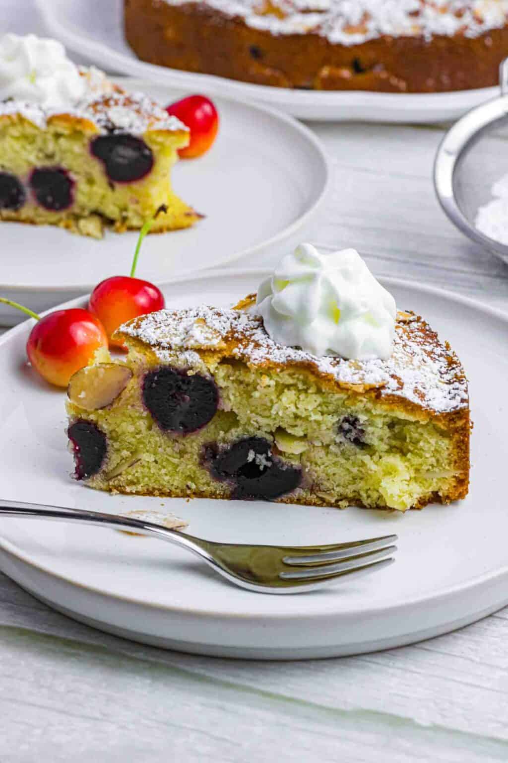 A piece of cherry almond cake on a white plate with whipping cream on top and 2 red cherries on the side.
