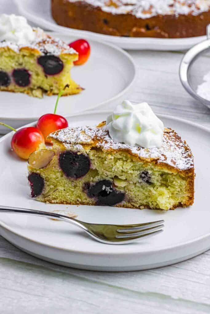A piece of cherry almond cake on a white plate with whipping cream on top and 2 red cherries on the side.