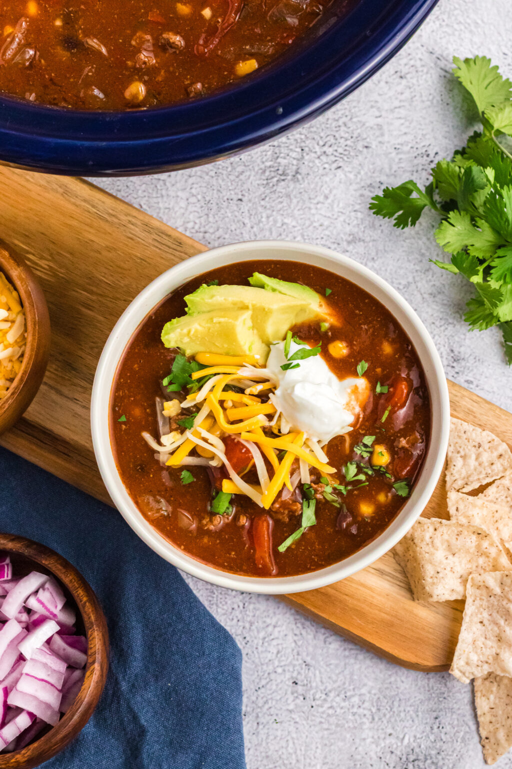 bowl of chili on a wooden board decorated with sour cream, shredded cheese, avocado slices and cilantro