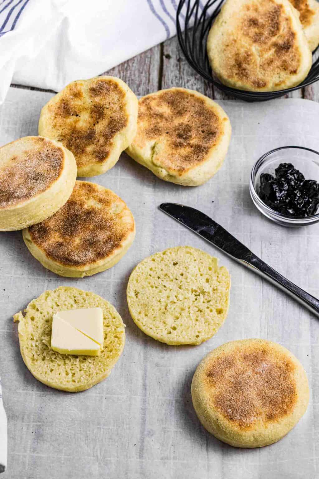 English Muffins on a breakfast table. One is cut open and has some butter on it.