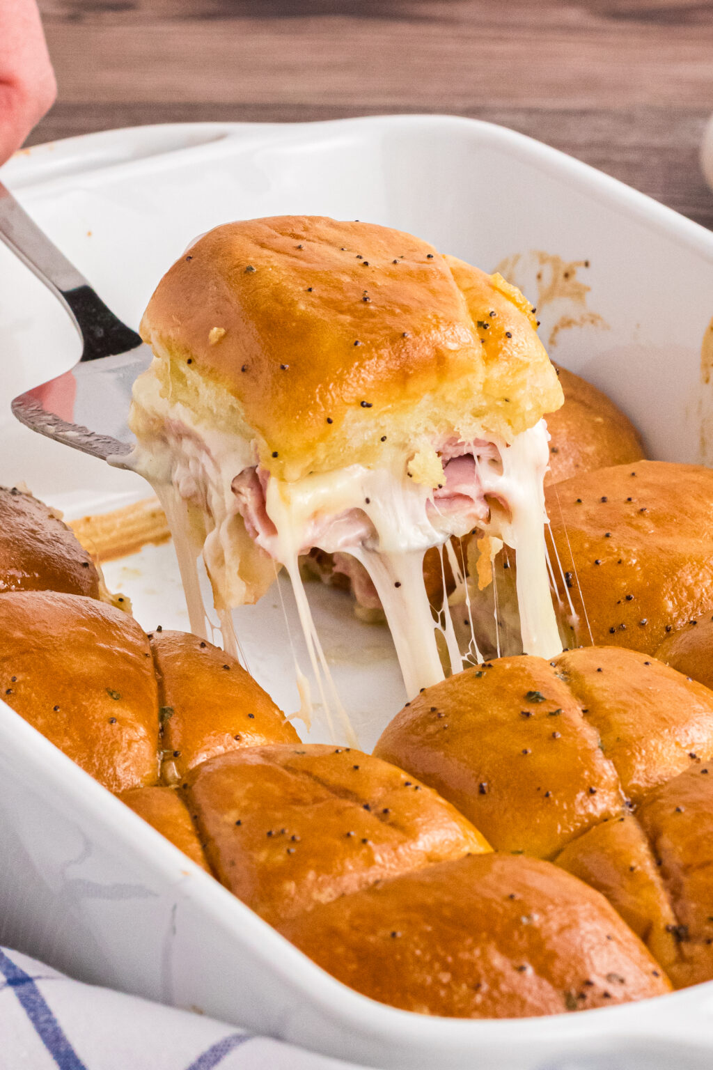 One of the Ham and Cheese sliders is being lifted out of the casserole featuring a stringy melty hot slider