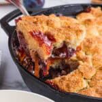 a big scoop of plum cobbler being lifted out of the black cast iron pan.