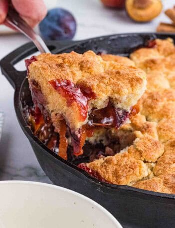 a big scoop of plum cobbler being lifted out of the black cast iron pan.