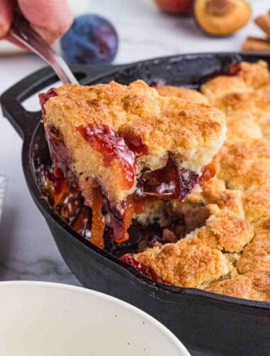 a big scoop of plum cobbler being lifted out of the black cast iron pan.
