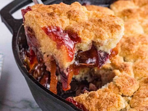 a big scoop of plum cobbler being lifted out of the black cast iron pan.