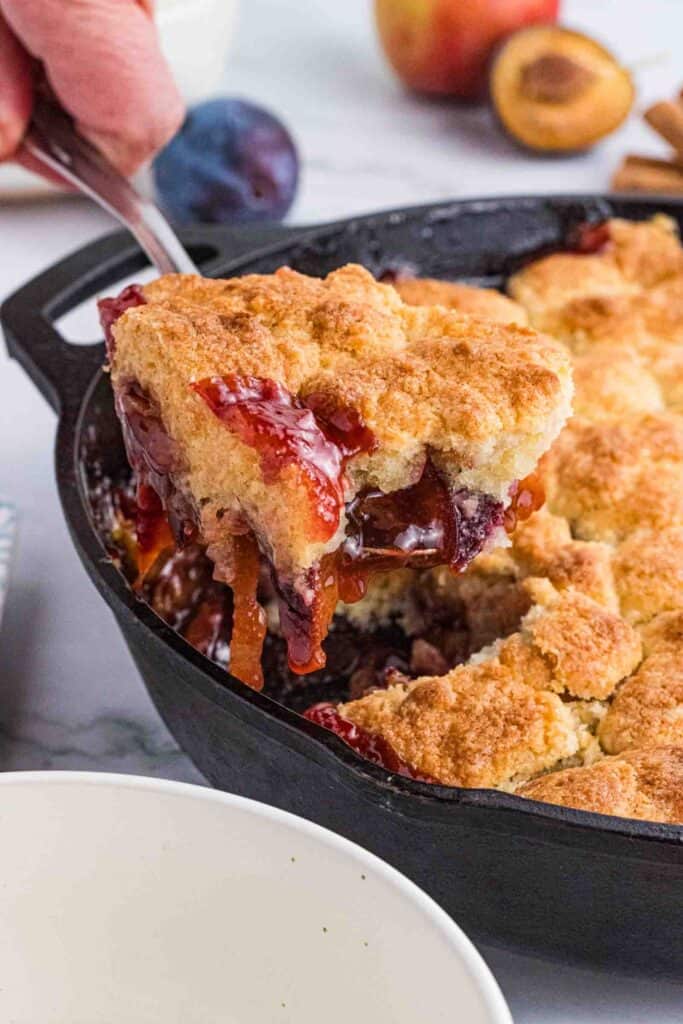 a big scoop of plum cobbler being lifted out of the black cast iron pan.
