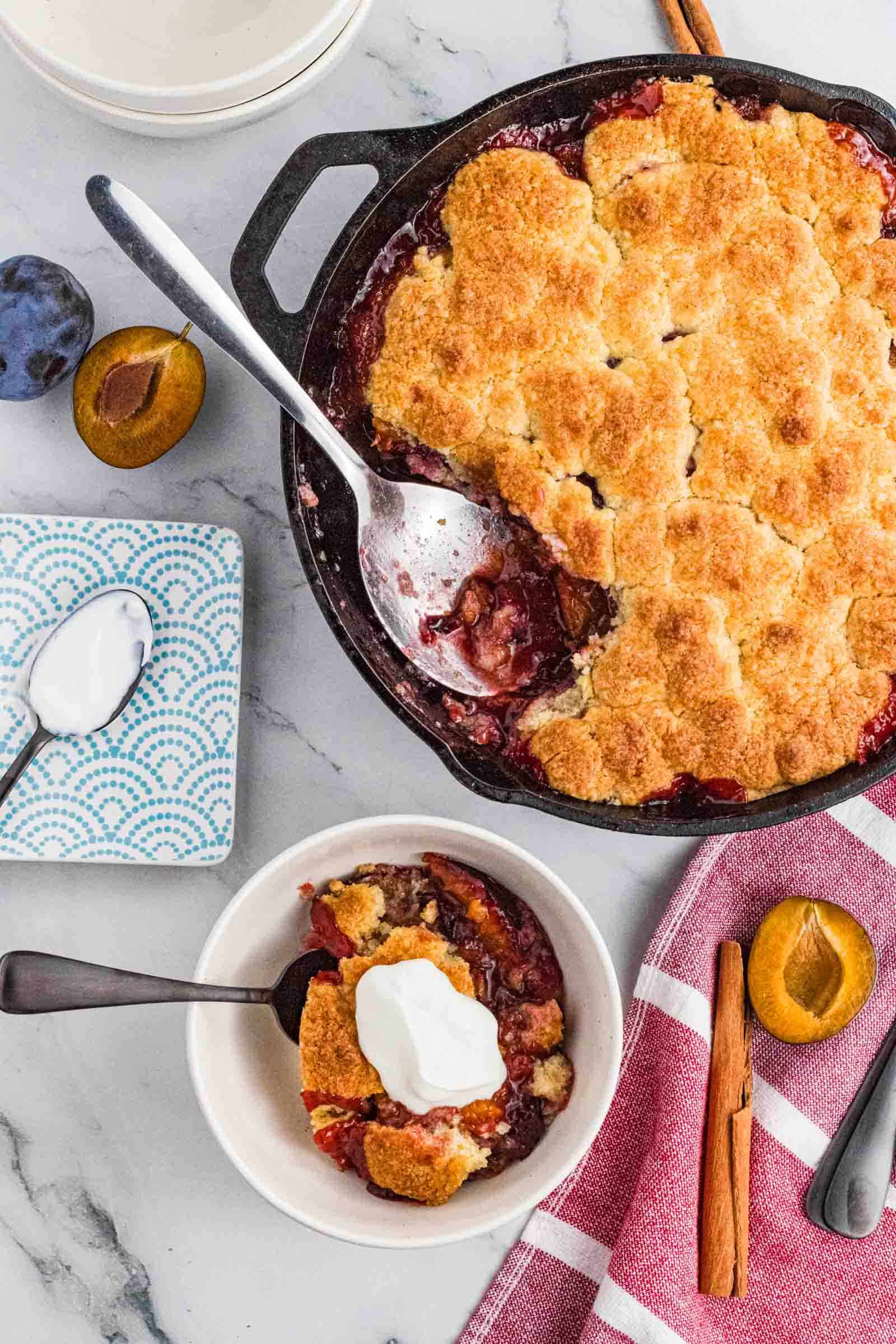 plum cobbler in cast iron skillet pan with some scoops in the white bowl topped with whipping cream