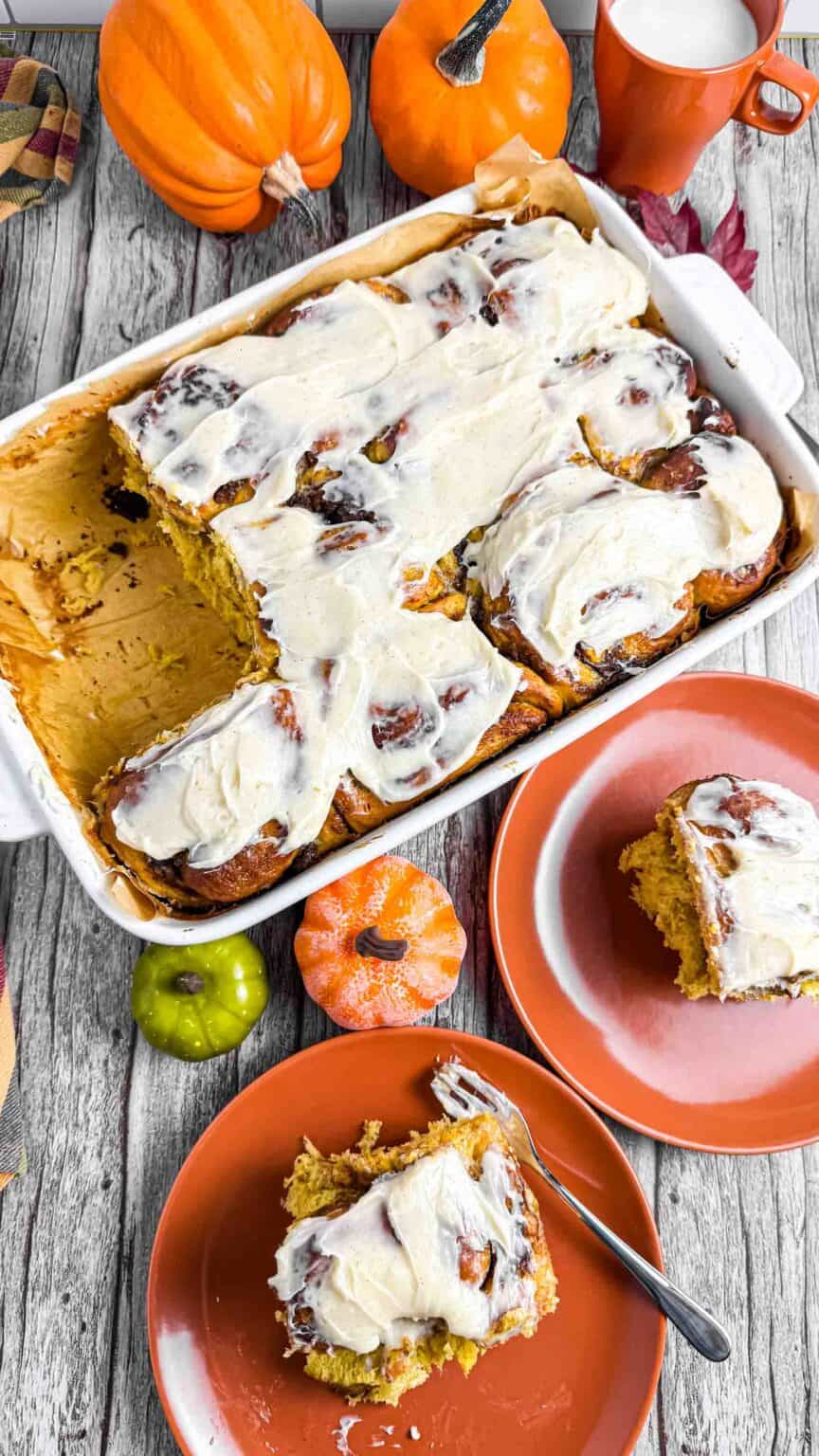 table decorated with pumpkin cinnamon rolls in the baking tray as well as two plates with each a roll on it.
