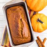 fresh baked pumpkin bread in tin surrounded by 2 pumpkins and cinnamon sticks