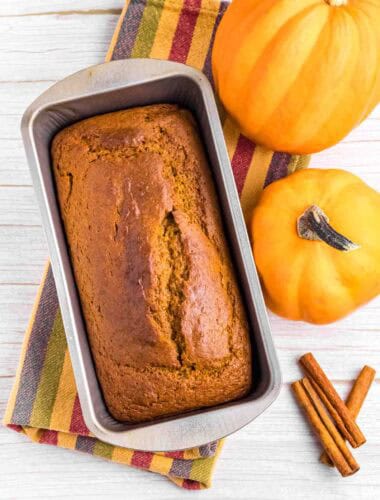 fresh baked pumpkin bread in tin surrounded by 2 pumpkins and cinnamon sticks