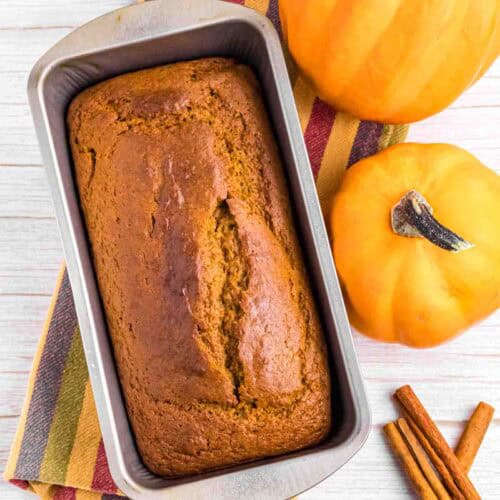 fresh baked pumpkin bread in tin surrounded by 2 pumpkins and cinnamon sticks