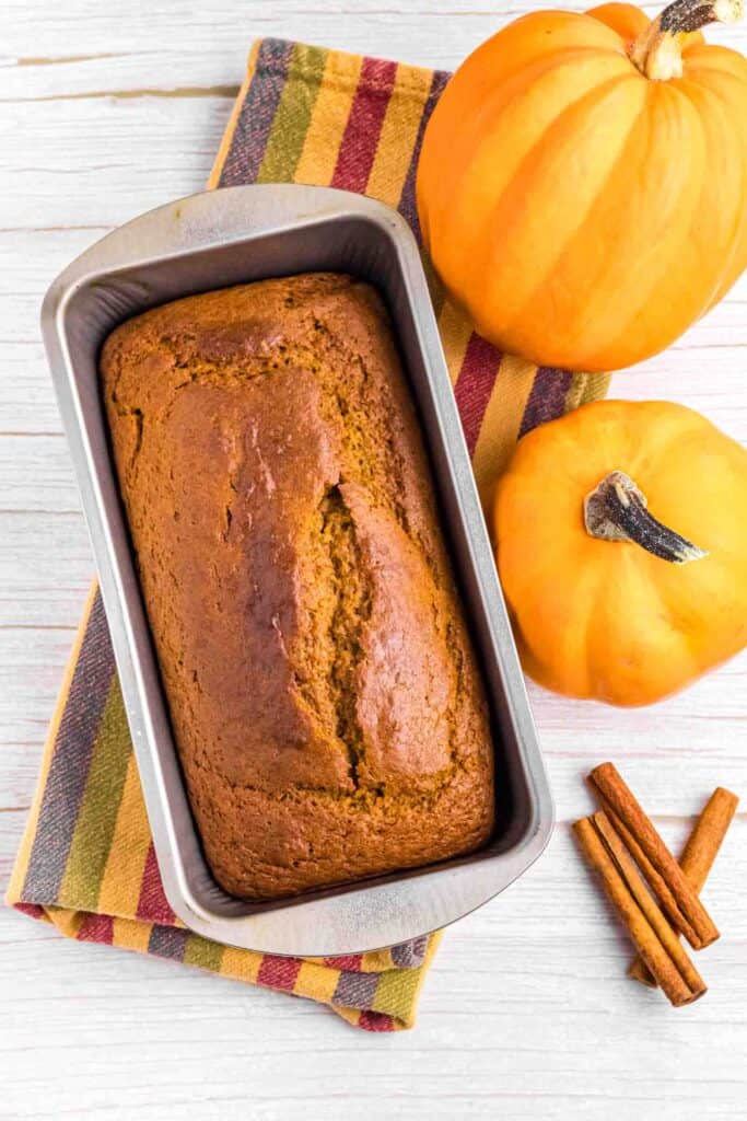 fresh baked pumpkin bread in tin surrounded by 2 pumpkins and cinnamon sticks