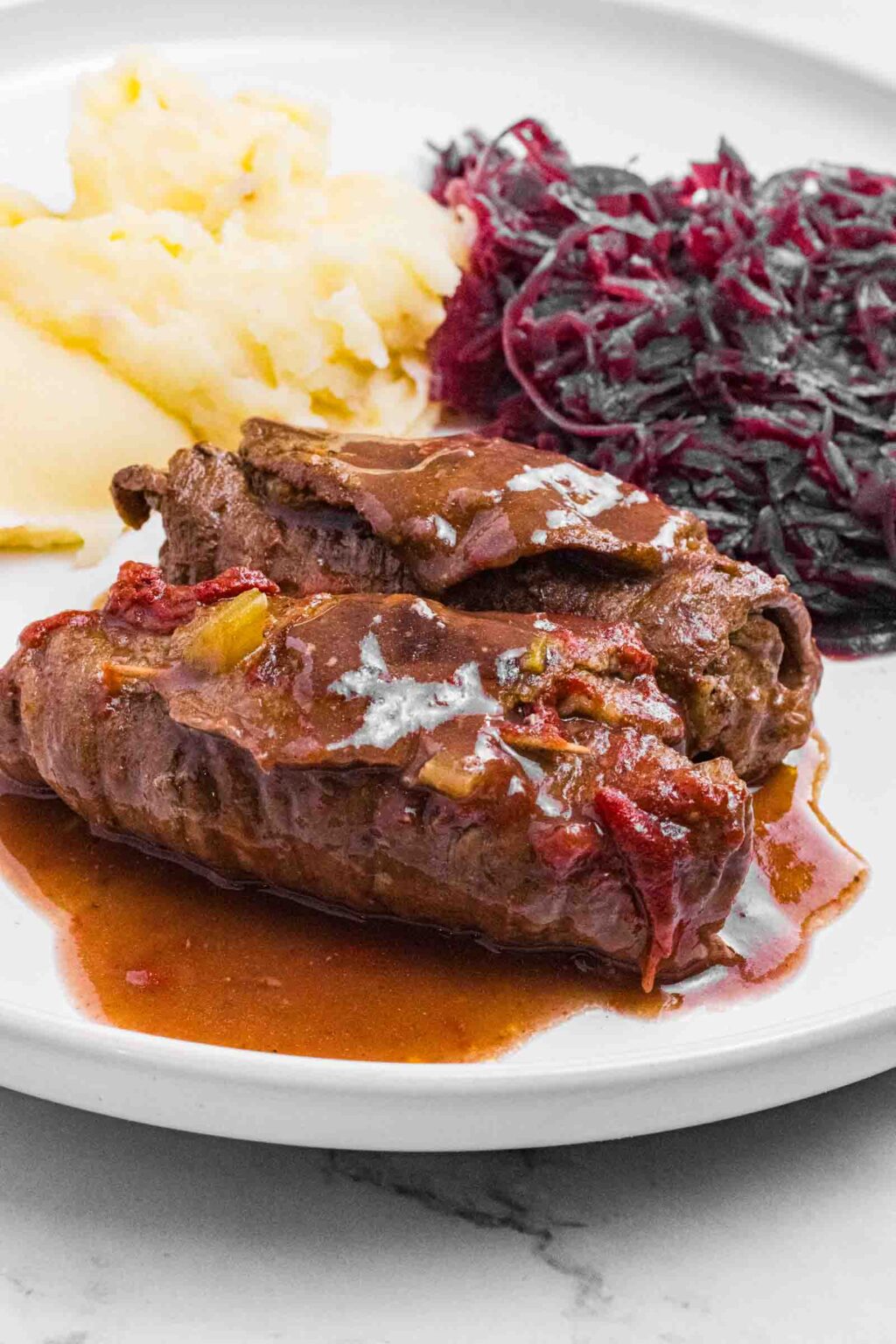 2 German Beef Rouladen on a white plate smothered with gravy and tucked in between mashed potatoes and red cabbage.
