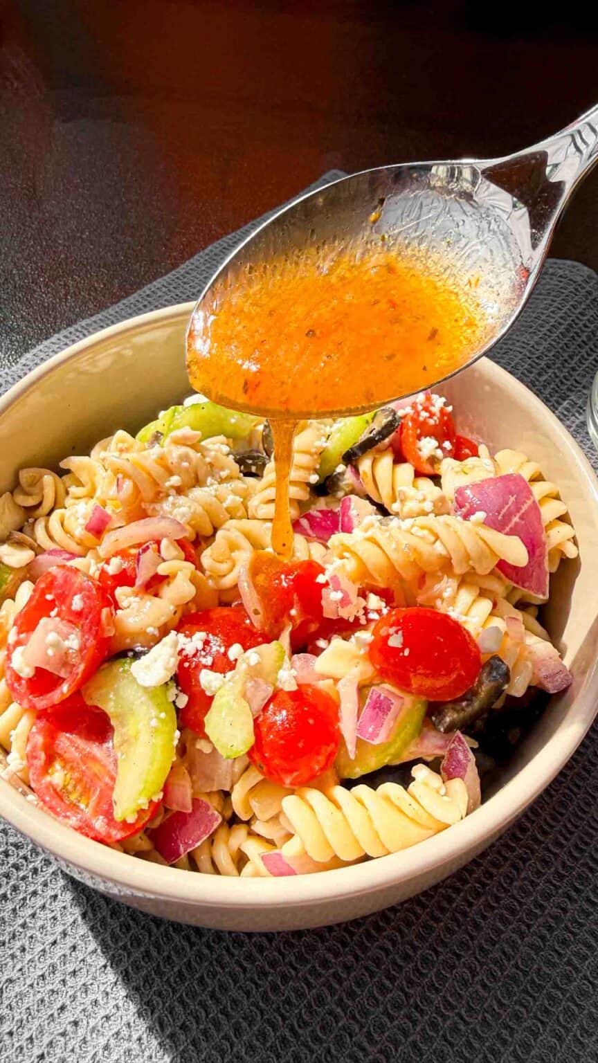 Italian dressing being drizzled over a bowl of mediterranean pasta salad