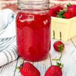 fresh strawberry sauce in a glass with 2 strawberries laying in front and a yellow strawberry basket behind it