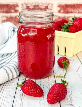 fresh strawberry sauce in a glass with 2 strawberries laying in front and a yellow strawberry basket behind it