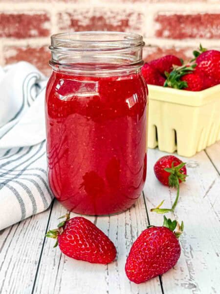 fresh strawberry sauce in a glass with 2 strawberries laying in front and a yellow strawberry basket behind it